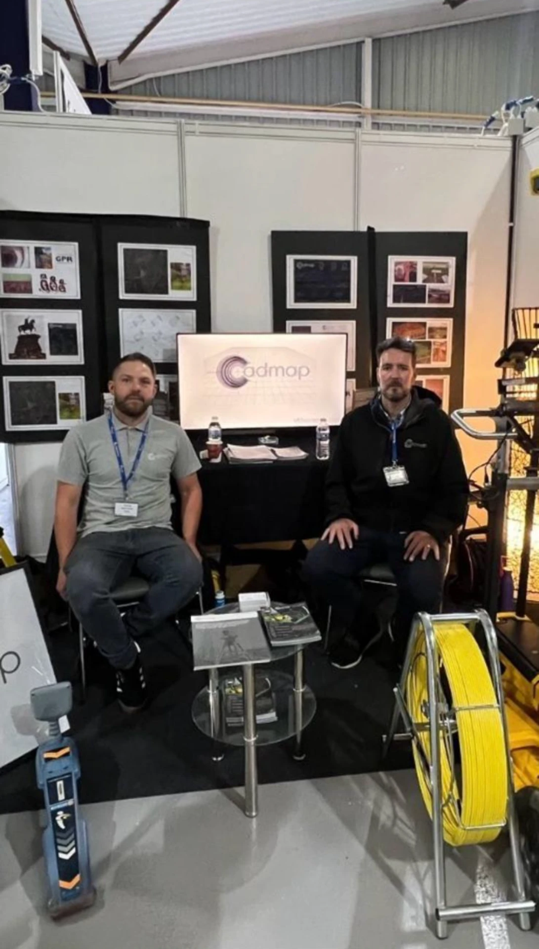 Two Cadmap surveyors seated at their exhibition stand with survey drawings, a total station and GPR equipment