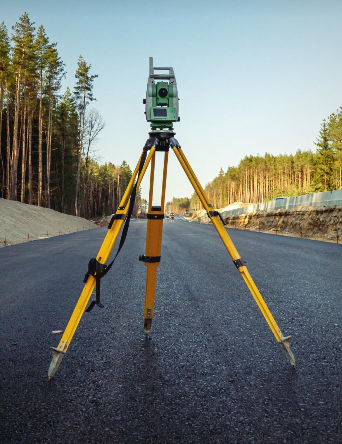 Total station on a yellow tripod on a newly surfaced road through woodland