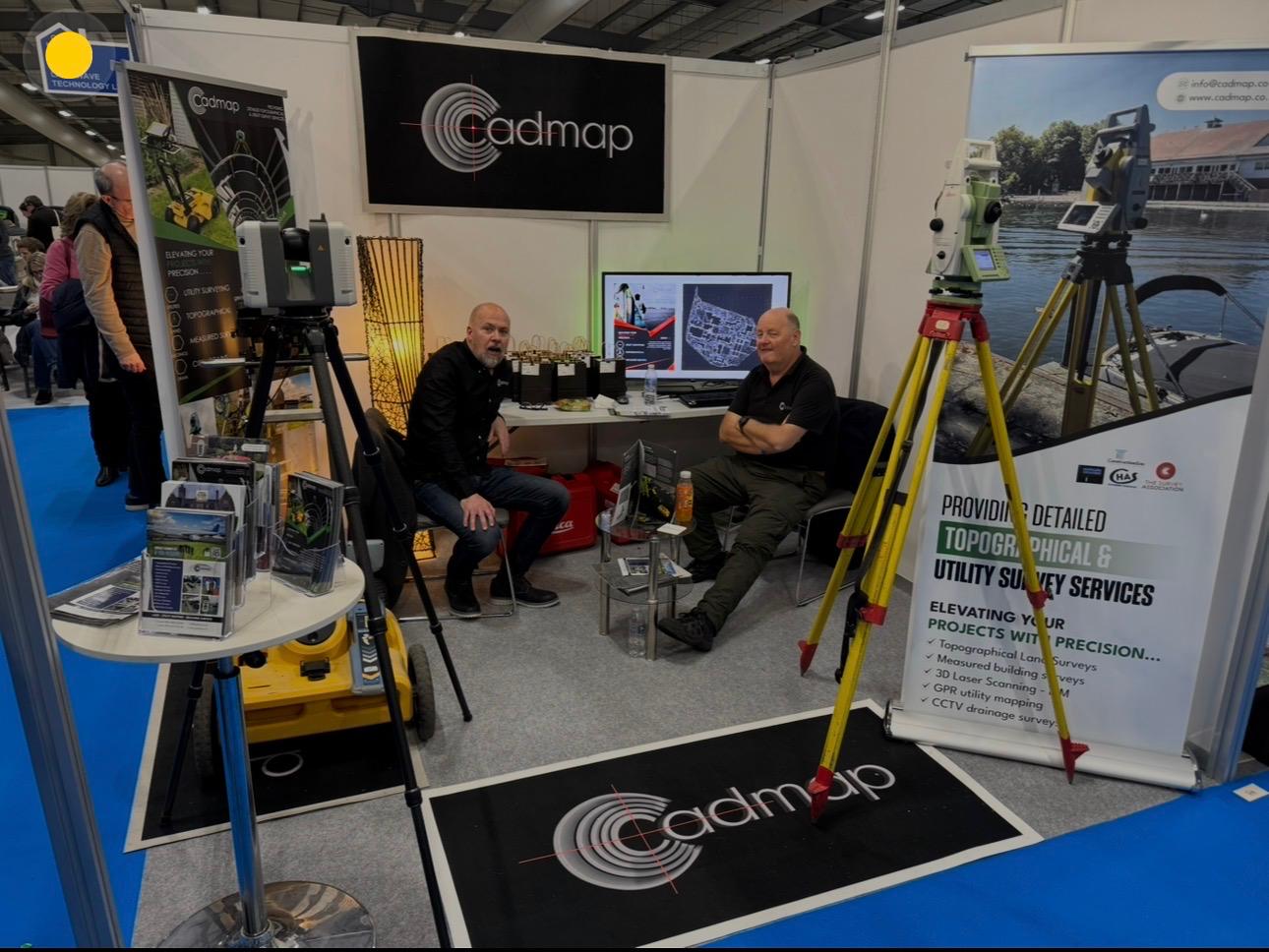 Two men seated at a CADMAP exhibition stand with surveying instruments and display banners
